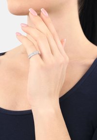 Un primer plano de una mano con un anillo de diamantes, los dedos tocando suavemente la barbilla, mostrando unas uñas bien cuidadas contra un fondo de ropa oscura.