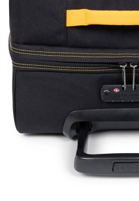 Musta kangasmatkalaukku, jossa on keltaiset korostukset ja ompeleet, ulosvedettävä kahva ja sivulla yhdistelmälukko.