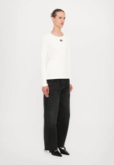 White long-sleeve ribbed top featuring a small black logo on the chest, paired with loose black denim jeans and pointed black shoes.