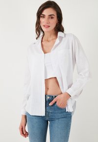 White, long-sleeve button-up shirt with a pocket, relaxed fit, lightweight fabric, and subtle texture, paired with blue jeans.