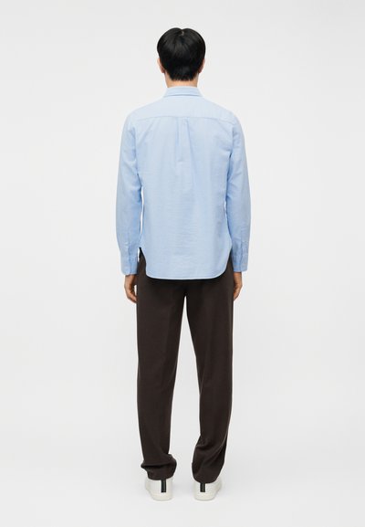 Mann, der ein hellblaues Hemd mit langen Ärmeln, dunkelbraune Hose und weiße Turnschuhe trägt, steht mit dem Rücken zur Kamera vor einem schlichten weißen Hintergrund.