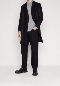 Cappotto nero doppiopetto su una camicia a righe, abbinato a pantaloni neri e scarpe stringate nere. Tessuto liscio con design strutturato.