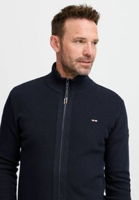 Jūrų laivyno zip-up megztinis su aukštu apykakle, tekstūrinio mezgimo raštu ir subtiliu logotipo akcentu. Turi užtrauktuką su auksinėmis detalėmis.
