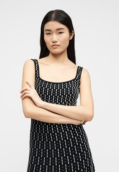 Jeune femme aux longs cheveux foncés portant une robe noire sans manches avec des lignes blanches pointillées verticales, les bras croisés, expression neutre, fond uni.