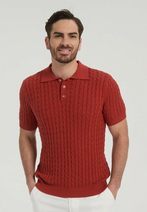Homme portant un polo en maille torsadée à manches courtes couleur rouille avec un pantalon blanc, debout les mains dans les poches devant un fond uni.