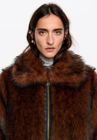 Brązowy sztuczny futro z szerokim kołnierzem, zapięciem na zamek błyskawiczny oraz paskowymi detalami, charakteryzujący się miękką teksturą i błyszczącym wykończeniem.