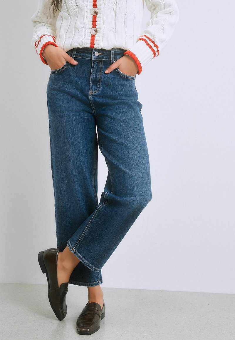 Persoon die een blauwe jeans met hoge taille, een witte gebreide trui met rode biezen en donkerbruine loafers draagt, staande met de handen in de zakken.