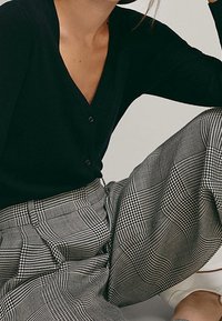 Cardigan noir côtelé avec col en V et fermeture à boutons, assorti à un pantalon à jambes larges avec un motif à carreaux noir et blanc.