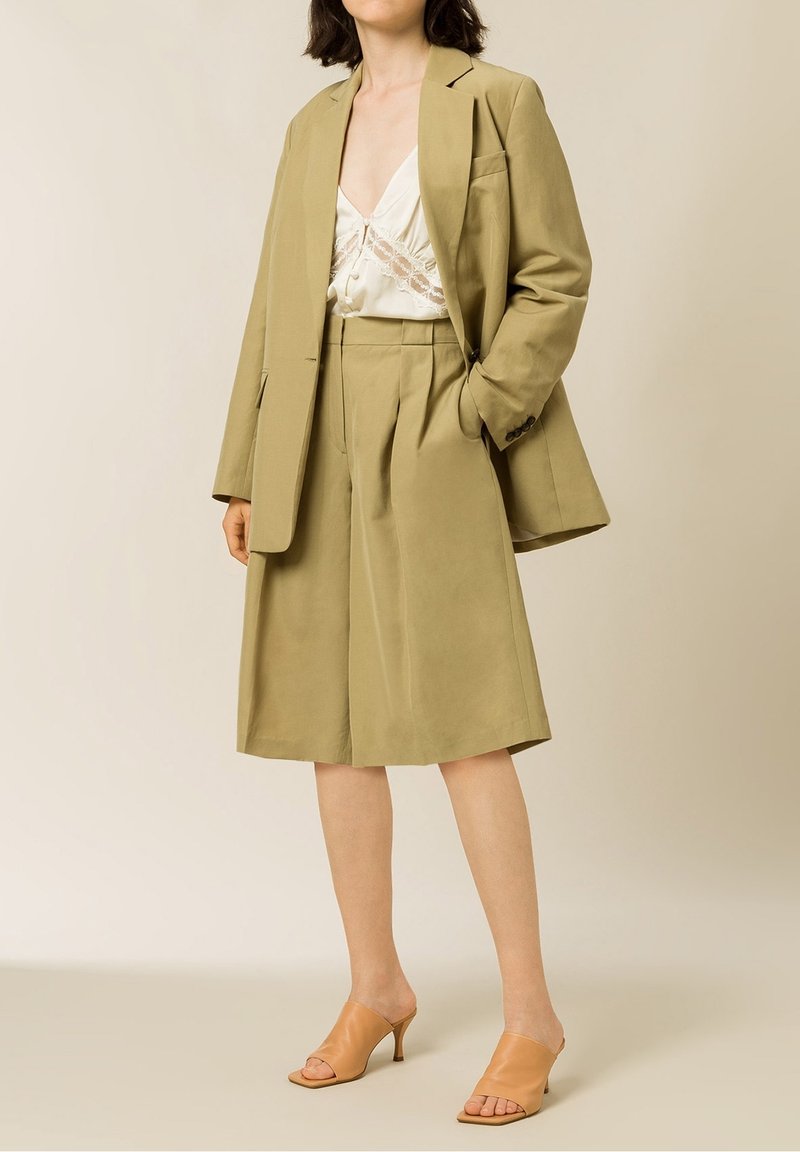 Femme portant un blazer beige ample et un short assorti jusqu’aux genoux, avec un chemisier blanc bordé de dentelle et des mules à talons couleur fauve, debout avec une main dans la poche.