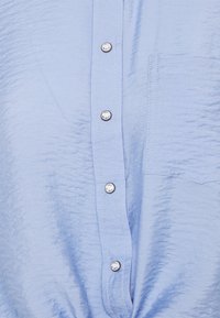 Ljusblå skjorta med knappar av krinklat tyg, som har silverfärgade knappar och en bröstficka på vänster sida. Slät textur.