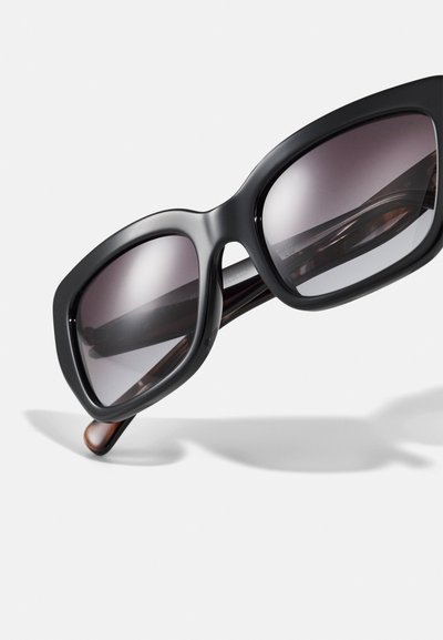 Mustat yli koko aurinkolasit tummilla gradienteilla varustetuilla linsseillä ja sileällä matalla kehyksellä. Ruskeat sävyiset templesit, joissa on kiiltävä pinta.