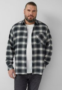 Camicia di flanella a quadri in bianco e nero con maniche lunghe, chiusura frontale con bottoni e una tasca sul petto, indossata sopra una maglietta bianca.