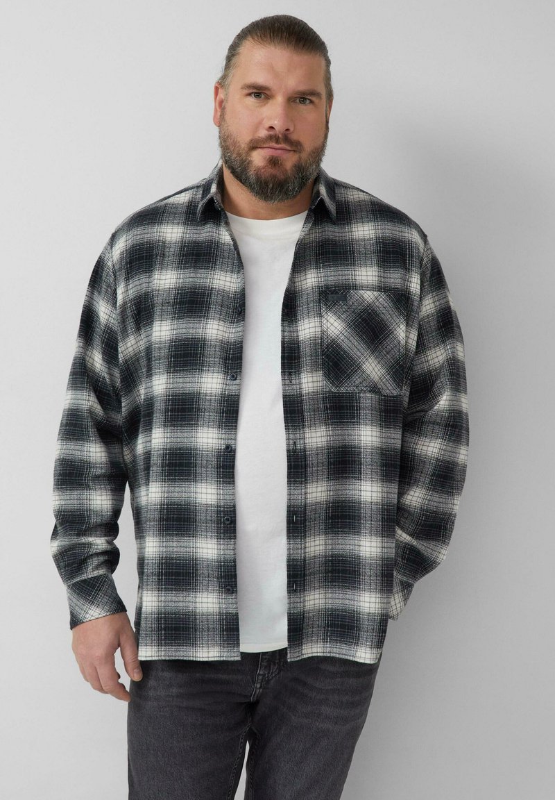 Camicia di flanella a quadri in bianco e nero con maniche lunghe, chiusura frontale con bottoni e una tasca sul petto, indossata sopra una maglietta bianca.