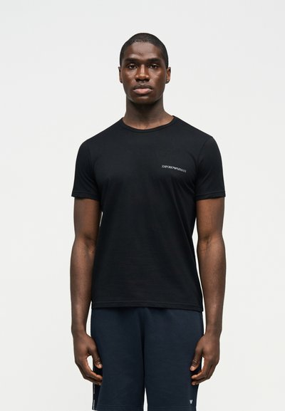 Άνδρας που στέκεται στραμμένος προς τα εμπρός, φορώντας μαύρο κοντομάνικο μπλουζάκι με μικρό λογότυπο και σκούρο σορτς, μπροστά σε απλό φόντο.