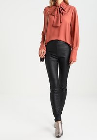 Roestkleurige blouse met een strikje bij de kraag, gecombineerd met zwarte glanzende leggings en zilveren enkellaarsjes. Soepele stof en aansluitend ontwerp.