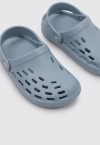 Helleblaue Kunststoff-Clogs mit durchbrochenem Design, runder Zehenpartie und Rückenschnalle. Mit einem Logo-Button auf jeder Seite. Glatte Textur.