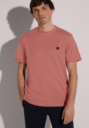 Uomo che indossa una maglietta a maniche corte color corallo smorzato con un piccolo logo circolare nero sul petto, abbinata a pantaloni scuri, in piedi davanti a uno sfondo chiaro.