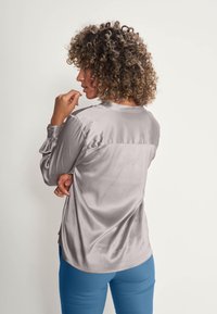 Blouse grise en soie avec un col rond, des manches longues plissées et un ourlet arrondi, associée à un pantalon bleu ajusté. Détail discret du logo au dos.