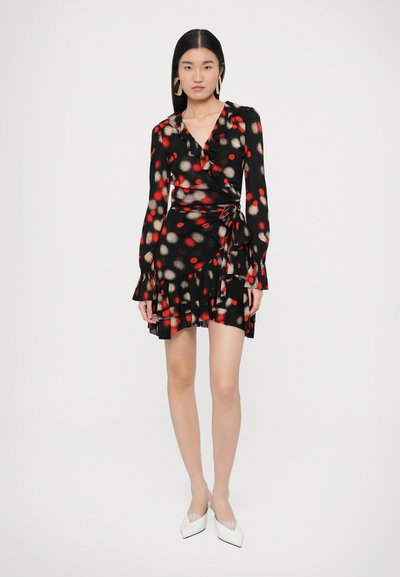 Μαύρο φόρεμα τύπου wrap με κόκκινες και μπεζ πουά, μακριά μανίκια, βολάν στο τελείωμα και ζώνη στη μέση. Φορεμένο με λευκά μυτερά παπούτσια.