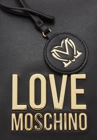 Superficie in pelle nera testurizzata con lettering dorato "LOVE MOSCHINO" e un'etichetta rotonda con un logo "M" a forma di cuore dorato, attaccata da una cinghia.