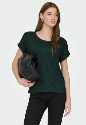 Jeune femme aux longs cheveux bruns porte un haut vert foncé à manches courtes et un pantalon noir, tenant un grand sac à main en cuir noir à la hauteur de la taille.