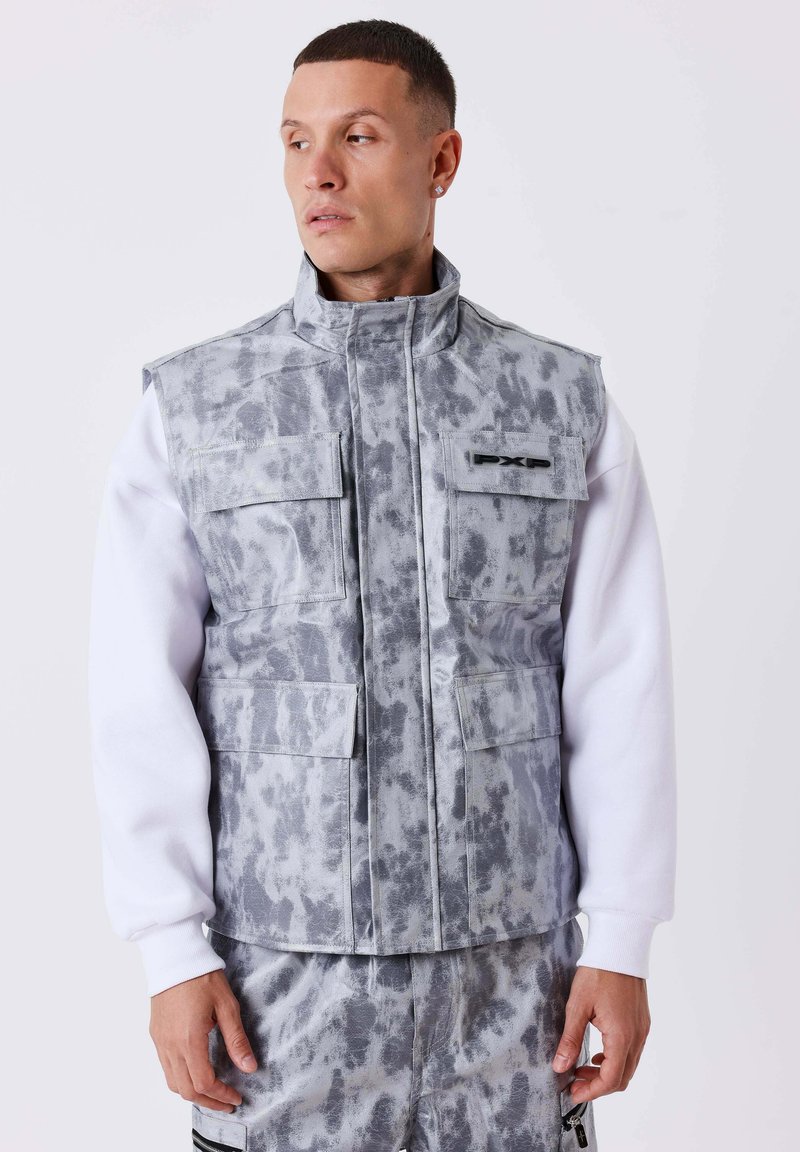 Homme portant un gilet utilitaire camouflage gris avec plusieurs poches par-dessus une chemise blanche à manches longues, regardant sur le côté sur un fond uni.