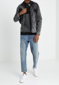 Personne portant une veste en jean noire délavée, un sweat-shirt noir et gris, un jean bleu clair, des baskets blanches, et tenant un sac bandoulière noir.