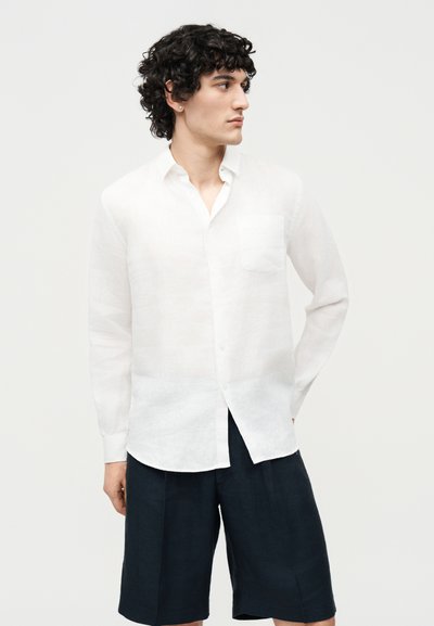 Άνδρας με σγουρά μαλλιά, φορώντας λευκό πουκάμισο με κουμπιά και μακρυά μανίκια και μαύρα σορτς μέχρι το γόνατο, κοιτάζει προς τα αριστερά του.