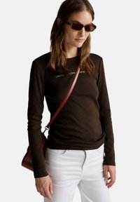 Ciemnobrązowa bawełniana koszula z długim rękawem z napisem "UNITED COLORS OF BENETTON". Połączona z białymi dżinsami i małą brązową torebką. Brązowe okulary przeciwsłoneczne.