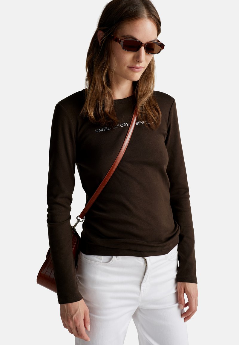 Ciemnobrązowa bawełniana koszula z długim rękawem z napisem "UNITED COLORS OF BENETTON". Połączona z białymi dżinsami i małą brązową torebką. Brązowe okulary przeciwsłoneczne.