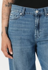 I jeans in denim blu presentano una vestibilità a vita media, un design a cinque tasche e una chiusura con zip e bottone metallico. Il tessuto ha una texture liscia con una leggera scoloritura.