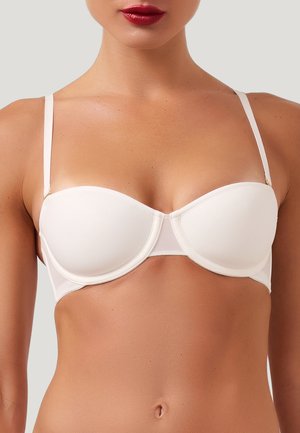 Kvinde iført en hvid stropløs bh med tynde justerbare stropper, viser den øverste del af overkroppen og rød læbestift på neutral baggrund.