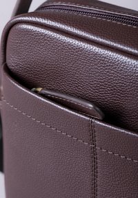 Brown leather bag with a textured finish, featuring a zippered closure and a side pocket with a curved handle. Stitching is visible along the edges.