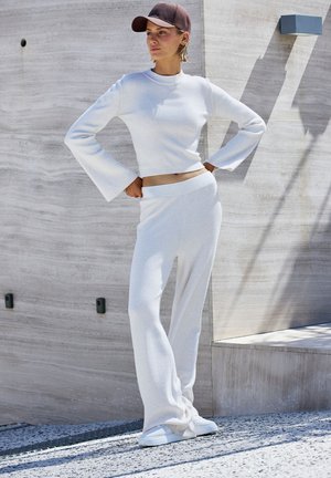 Weißes, geripptes Strick-Zweiteiler-Outfit mit kurzem Langarmoberteil und ausgestellten Ärmeln sowie passenden weit geschnittenen Hosen, vor einem strukturierten Hintergrund.