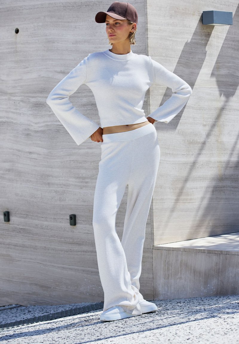 Balta raižyta megzta dviejų dalių apranga, kurioje yra trumpintas, ilgomis rankovėmis viršus su platėjančiomis rankovėmis ir derančios platėjančios kelnės, fone matyti tekstūruotas fonas.