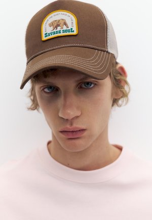 Giovane con capelli chiari e ricci, che indossa un berretto marrone e bianco con una toppa a forma di orso "Savage Soul" e una maglietta rosa pallido su sfondo bianco.