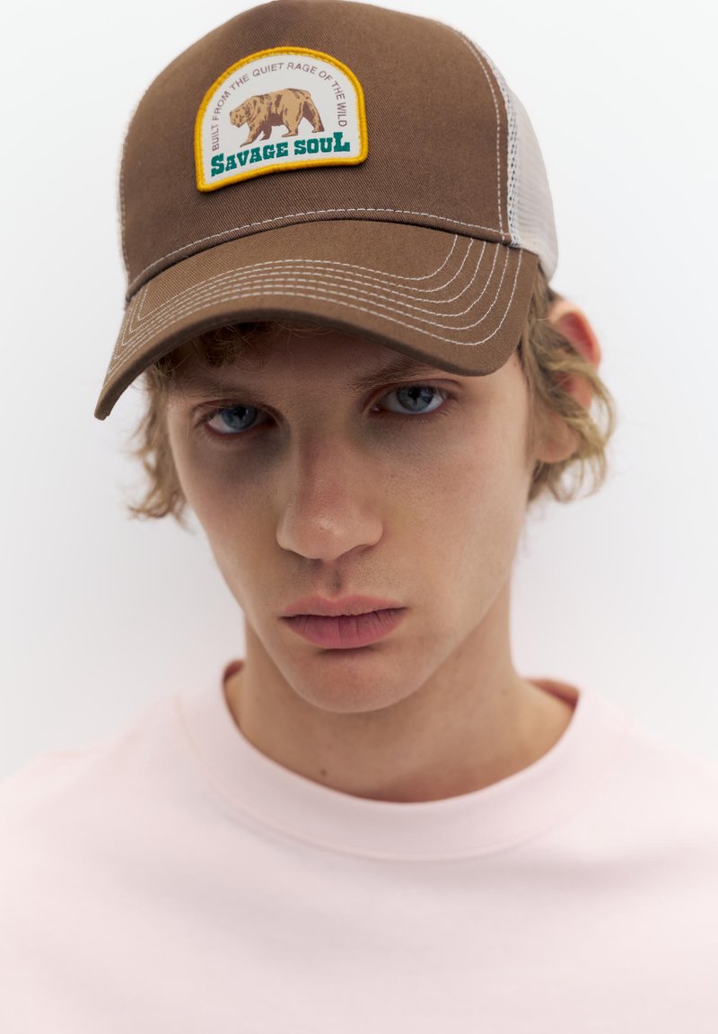 Jeune personne aux cheveux bouclés clairs, portant une casquette marron et blanche avec un écusson ours "Savage Soul" et une chemise rose pâle sur fond blanc.