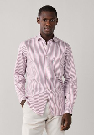 Hombre con camisa blanca de manga larga con rayas verticales rojas y pantalones de color claro, de pie frente a un fondo liso.