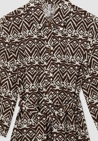 Brun och krämfärgad mönstrad skjortklänning med krage, knappstängning framtill, långa ärmar och knytbälte i midjan. Har geometriska mönster.