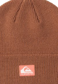 Bonnet en tricot marron avec texture côtelée, comportant un patch logo rose. Forme effilée avec un revers replié à la base.
