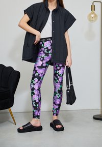 Giacca nera trapuntata senza maniche, t-shirt bianca, leggings floreali con motivo viola e verde, sandali neri flat, in mano una borsa nera.