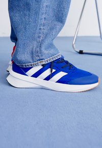 Ténis atléticos azuis com riscas brancas, parte superior texturizada e sola de borracha lisa. Usados com jeans azul claro.