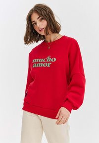 Czerwona bluza z napisem "mucho amor" w zielonym kolorze. Charakteryzuje się luźnym krojem, ściągaczami przy rękawach i dolnym brzegu, wykonana jest z miękkiego materiału. Subtelna metka z logo umieszczona u dołu.