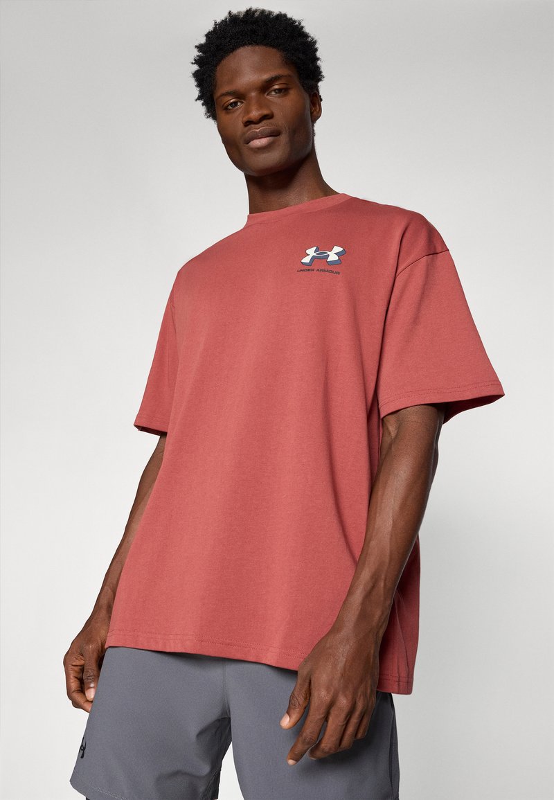 Camiseta de algodón roja con mangas cortas y cuello redondo, que presenta un logo de Under Armour en azul y blanco en el pecho. Corte casual.