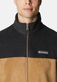 Giacca in pile con una parte superiore nera e una parte inferiore beige, con chiusura a zip e logo Columbia sul lato sinistro.