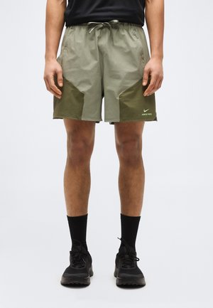 Uomo che indossa pantaloncini Nike Pro verde chiaro e verde scuro, calzini neri e scarpe da ginnastica nere, in piedi davanti a uno sfondo semplice.