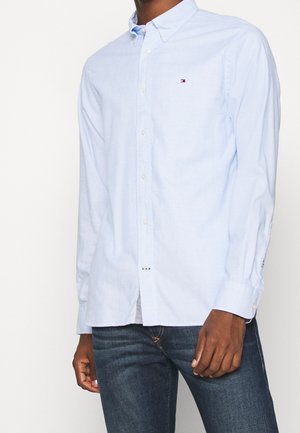 Mann trägt hellblaues Hemd mit kleinen Logo auf der Brust und dunkelblaue Jeans, steht vor einem schlichten weißen Hintergrund.