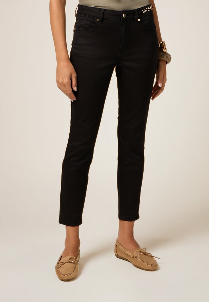 Jeans negros ajustados con cinco bolsillos, cremallera frontal y herrajes metálicos, combinados con zapatos estilo mocasín en tono marrón claro. Textura de tela suave.