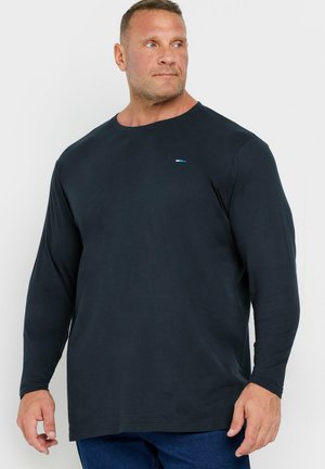 Man with short hair wearing a plain black long-sleeve shirt and blue jeans, looking to his right against a white background.