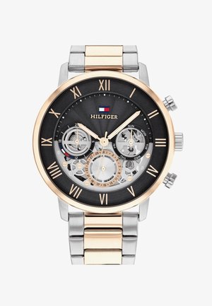 Zegarek chronograficzny z czarną tarczą, obudowa z nierdzewnej stali w kolorze złotym i srebrnym, cyfry rzymskie oraz widoczne wewnętrzne mechanizmy. Posiada wskaźniki dnia/miesiąca.